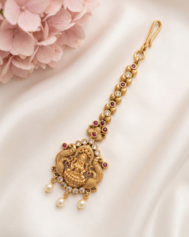 An ornate gold-toned traditional Indian jewelry piece, likely a Golu Kara or Gopalakrishnanjali pendant, featuring a detailed Ganesha motif, red stones, and white pearls, displayed on a white satin fabric with pink flowers in the corner.
