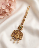 An ornate gold-toned traditional Indian jhumka earring with a detailed Ganesha pendant, set against a draped white fabric background with pink flowers in the corner.