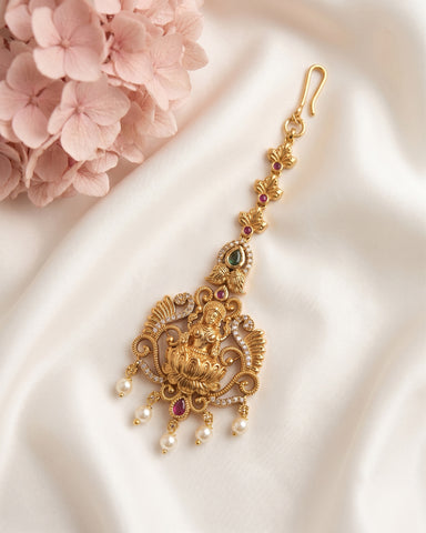 An ornate gold-toned traditional Indian earring with detailed filigree, small red and green stones, and dangling pearl-like beads, displayed on white satin fabric with pink flowers in the corner.