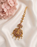 An ornate gold-toned traditional Indian maang tikka with red gemstone accents and hanging pearl beads, displayed on draped white satin fabric next to pink flowers.