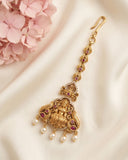 An ornate gold-toned traditional Indian earring featuring a detailed Ganesha motif, accented with small red gemstones and white pearl drops, displayed on a draped cream fabric with pink flowers in the corner.