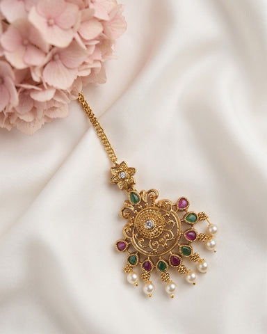 An ornate gold-toned Indian maang tikka pendant with green and red gemstone accents and dangling pearl beads, displayed on a draped white satin fabric next to pink flowers.