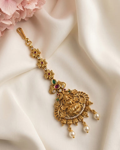 An ornate gold-toned traditional Indian jewelry piece, likely a maang tikka or pendant, decorated with small green and red stones and white pearls, displayed on draped white satin fabric with pink flowers in the corner.