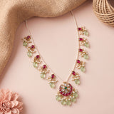 A gold-plated statement necklace with green glass beads, white faux pearls, and red stones, styled on a pink background with burlap and a flower.