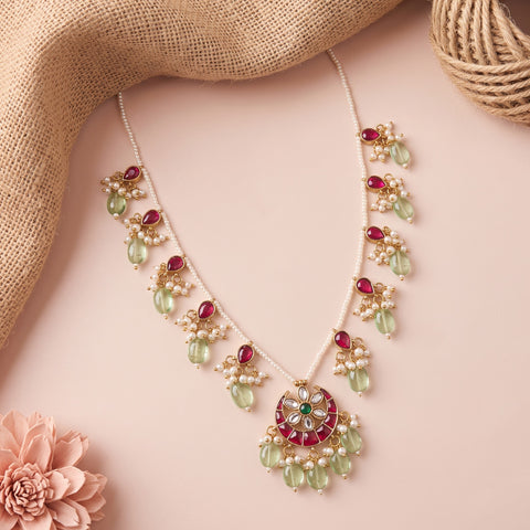 A gold-plated statement necklace with green glass beads, white faux pearls, and red stones, styled on a pink background with burlap and a flower.