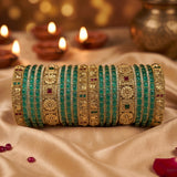 A stack of traditional Indian bangles (kara) featuring green stone-embellished bands alongside ornate gold-plated filigree bangles with small red and green stone accents, displayed on draped beige fabric with blurred diya candles in the background.