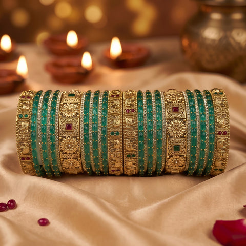 A stack of traditional Indian bangles (kara) featuring green stone-embellished bands alongside ornate gold-plated filigree bangles with small red and green stone accents, displayed on draped beige fabric with blurred diya candles in the background.