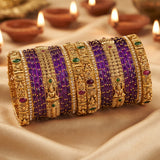 A set of traditional Indian bangles, featuring purple beaded bracelets paired with ornate gold-plated filigree bangles decorated with small green and red stones, arranged on draped beige fabric with blurred diya candles in the background.