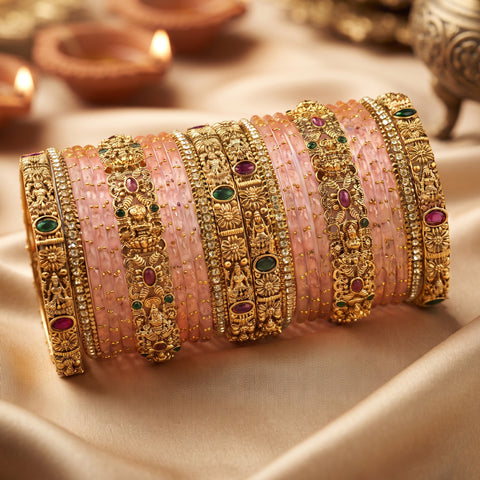 A stack of ornate Indian bangles, including pink stone-wrapped bangles and detailed gold-plated bangles with red and green stones, displayed on a beige satin fabric with blurred diya candles in the background.