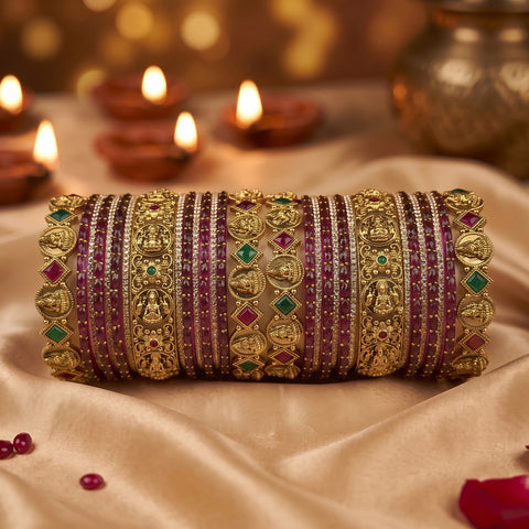 A set of 18 traditional Indian bangles (kara) made of gold-plated metal, decorated with rows of deep pinkish-red stones, small clear stones, and green and red gem accents. The bangles are arranged horizontally on draped beige satin fabric with blurred lit diya candles in the background.