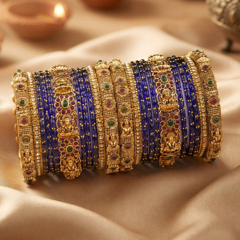 A stack of traditional Indian gold-plated bangles with intricate filigree work, set with small red, green, and blue stones, alongside solid royal blue bangles, displayed on draped beige satin fabric.