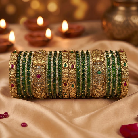A stack of traditional Indian bangles (kara bracelets) made of gold-plated metal with green stone inlays and small red and green gemstone accents, displayed on a beige satin fabric with blurred diya candles in the background.