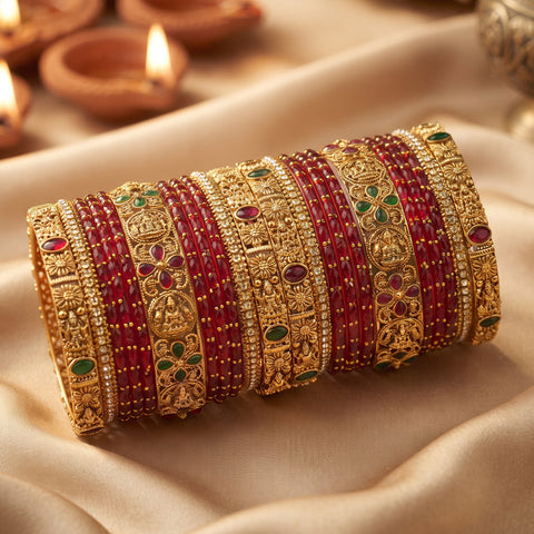 A stack of ornate Indian bangles (kara) featuring gold-plated filigree bands with red bead rows and small green and red stone accents, displayed on draped beige fabric with blurred diya lights in the background.