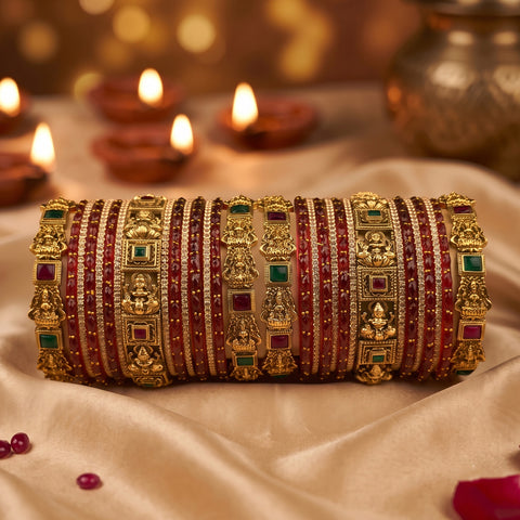 A stack of ornate gold bangles decorated with red and green stones, displayed on a beige satin fabric with blurred lit diya candles in the background.
