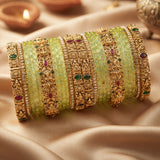 A stack of traditional Indian bangles, featuring light green stone-set bands alongside ornate gold-plated filigree bangles decorated with red and green stone accents, displayed on draped beige satin fabric.
