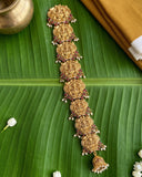 An ornate gold-toned traditional Indian jewelry piece, likely a necklace or pendant set, decorated with floral motifs and small red and white bead accents, displayed on a large green banana leaf.