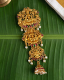 An ornate gold-toned traditional Indian jewelry piece featuring multiple deity motifs, red gemstone accents, and white pearl beads, displayed on a green banana leaf.