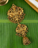 An ornate gold-toned traditional Indian jhumka earring with detailed deity carvings and dangling bells, displayed on a green banana leaf.