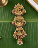 An ornate gold-toned traditional Indian jewelry piece with detailed carvings, red and green stone accents, and dangling pearl-like beads, displayed on a large green banana leaf.