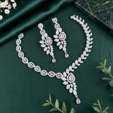 Silver-toned jewelry set with a necklace, matching earrings, and a curved bracelet, decorated with clear stones and pale pink teardrop accents, arranged on a dark green satin fabric with small floral and leaf props.