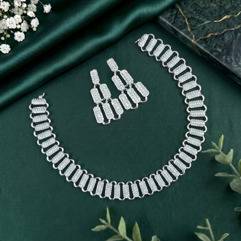 A silver-toned jewelry set featuring a necklace and matching earrings, both adorned with pavé-set clear stones in a repeating rectangular link pattern, arranged on a dark green satin fabric background with decorative floral and marble props.