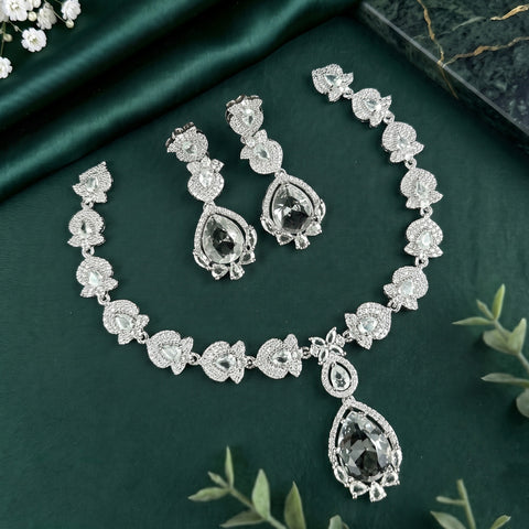 A silver-toned jewelry set featuring a necklace, matching earrings, and bracelet, decorated with clear rhinestones and large teardrop-shaped dark crystal pendants, arranged on a dark green satin fabric background.