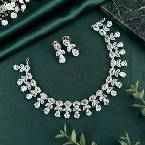 A silver-toned jewelry set featuring a necklace, bracelet, and matching earrings, all decorated with clear teardrop and oval stones in a floral leaf pattern, arranged on a dark green satin fabric background.