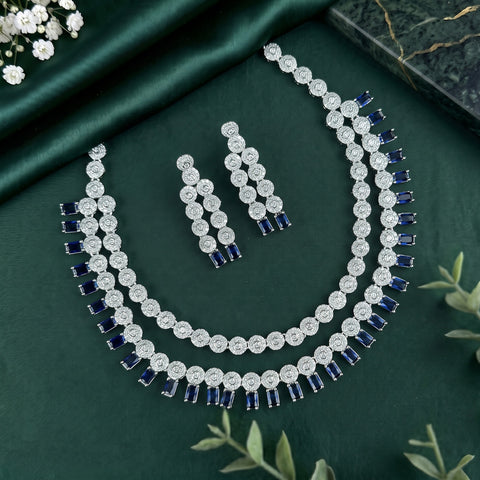 A silver-tone jewelry set featuring a double-strand necklace and matching drop earrings, embellished with round clear stones and rectangular dark blue stones, arranged on a dark green satin fabric background.