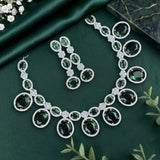 A jewelry set featuring a necklace and matching drop earrings with large oval dark green stones surrounded by silver-toned pavé settings, arranged on a dark green satin fabric with small white flowers and greenery accents.