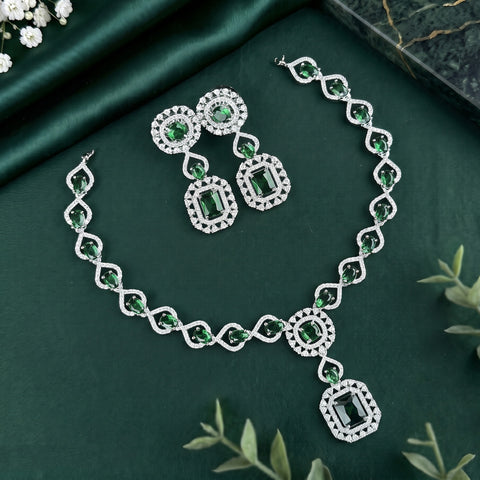 Silver-tone jewelry set with green emerald-cut stones, including a necklace, matching earrings, and a bracelet, arranged on a dark green satin fabric background with decorative leaves and flowers.