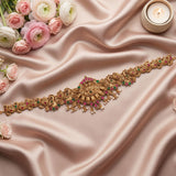 An ornate gold-toned traditional waist chain or belt with intricate floral and peacock motifs, embellished with pink and green stones and small pearl-like drops, laid out on draped blush satin fabric with pink and white flowers and a lit candle around it.