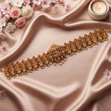 An ornate gold-toned traditional necklace with detailed deity motifs and small bead drops, arranged diagonally on draped champagne satin with pink flowers and a lit candle in the corners.