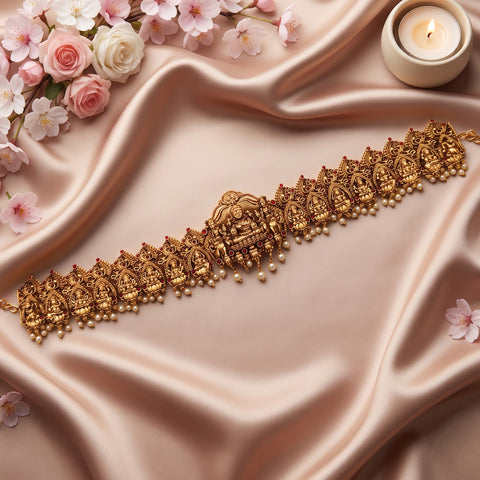 An ornate gold-toned traditional necklace with detailed deity motifs and small bead drops, arranged diagonally on draped champagne satin with pink flowers and a lit candle in the corners.