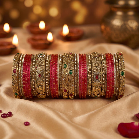 A set of traditional Indian bangles (kara) displayed horizontally on a beige satin fabric. The bangles are primarily red and gold, with some featuring green stone accents.