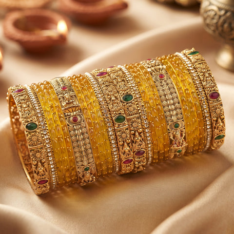 A stack of traditional Indian gold bangles with intricate floral engravings and small green, red, and pink stone accents, displayed on draped cream satin fabric.