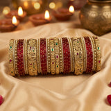 A set of traditional Indian bangles (kara) displayed in a stack. The bangles feature alternating rows of deep red beads and intricate gold-plated metal bands decorated with filigree, small stones, and tiny red and green accents.