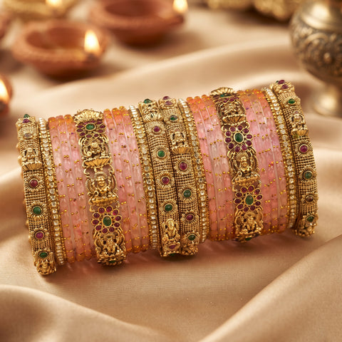 A stack of traditional Indian bangles, featuring pink stone-set bands alongside ornate gold-plated rings with deity motifs and green and red stone accents, displayed on draped beige fabric with blurred diya lights in the background.