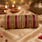 A stack of ornate Indian bangles (batches) featuring bright pinkish-red enamel stripes and intricate gold-plated detailing, displayed on a beige satin fabric with warm bokeh background lights.