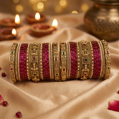 A stack of ornate Indian bangles (batches) featuring bright pinkish-red enamel stripes and intricate gold-plated detailing, displayed on a beige satin fabric with warm bokeh background lights.