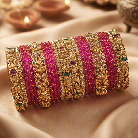 A set of traditional Indian bangles, including magenta pink beaded bracelets and ornate gold-plated filigree bangles with green and red stones, arranged on draped beige fabric with blurred diya candles in the background.