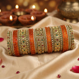 A stack of traditional Indian bangles (brocaches) made of gold and silver-toned metals, decorated with orange and red beaded stripes and small green and red stones, displayed on draped beige satin fabric with blurred diya candles in the background.