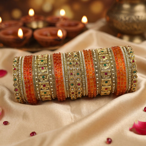 A stack of traditional Indian bangles (brocaches) made of gold and silver-toned metals, decorated with orange and red beaded stripes and small green and red stones, displayed on draped beige satin fabric with blurred diya candles in the background.