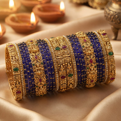 A stack of ornate gold bangles with blue, red, and green stone accents, displayed on draped satin fabric with warm background lights.