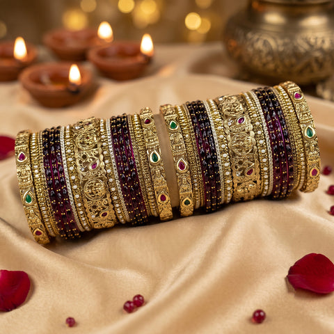 A set of ornate gold-toned bangles with red, green, and purple beaded accents, arranged on draped beige satin fabric with lit diya candles and rose petals in the background.