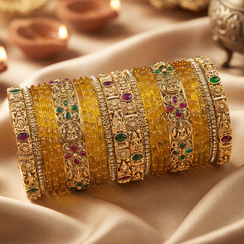 A stack of traditional Indian gold bangles (batches) decorated with yellow stones, green and pink gemstones, and silver-toned metal engravings, displayed on draped beige satin fabric with blurred diya candles in the background.