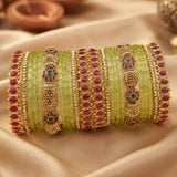 A set of traditional Indian bangles (kara) featuring light green stone-encrusted bands, gold-plated filigree, and rows of red and green gemstones, displayed on a beige satin fabric.