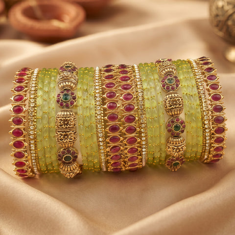 A set of traditional Indian bangles (kara) featuring light green stone-encrusted bands, gold-plated filigree, and rows of red and green gemstones, displayed on a beige satin fabric.
