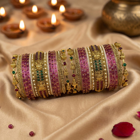 A set of traditional Indian bangles (batches) featuring pink stone inlays, gold-plated borders, and small green and red stone accents, displayed on a beige satin fabric with lit diya candles in the background.
