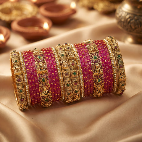 A stack of traditional Indian bangles, featuring bright pinkish-red striped bands alongside ornate gold-plated filigree bangles embellished with small green and red stones, displayed on draped beige fabric with blurred diya lamps in the background.