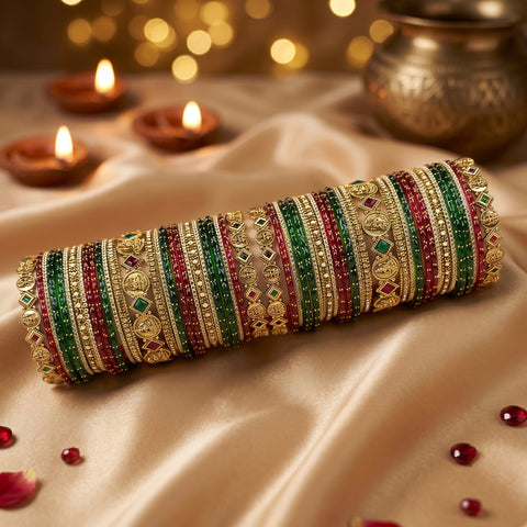 A stack of traditional Indian bangles (kara) made of gold-plated metal, decorated with green and red stones, displayed on a draped beige satin fabric with lit diya candles in the background.